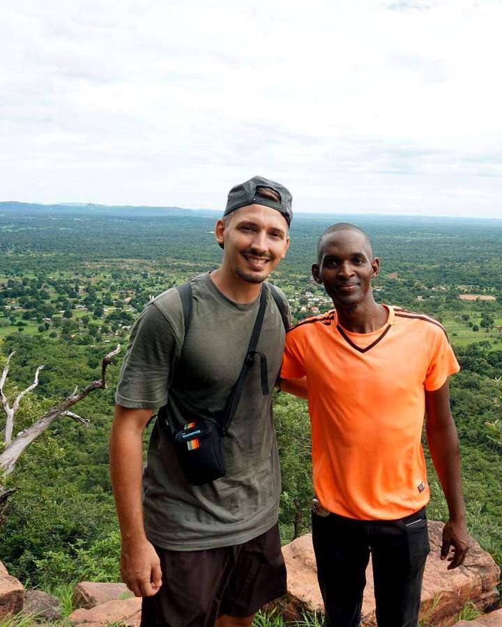 Bouba, con camiseta naranja, posa sonriente junto a un viajero en un mirador sobre el valle verde