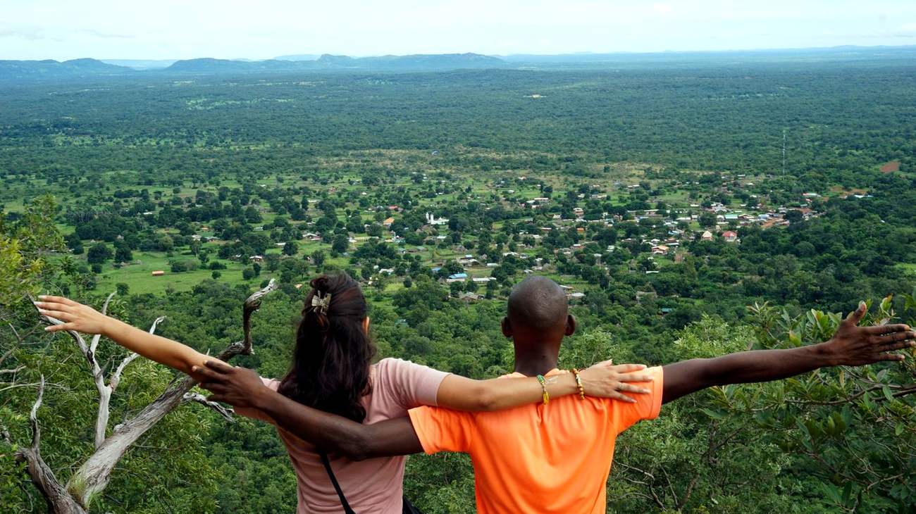 Bouba y una viajera, de espaldas y con los brazos abiertos, contemplan desde lo alto el valle verde y un pueblo entre los árboles